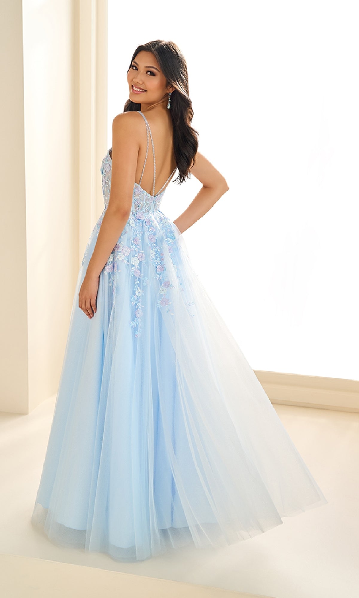 A woman in the Ellie Wilde EW36079, a light blue, floor-length prom dress with floral embroidery and a corset bodice, smiles over her shoulder in a softly lit room.